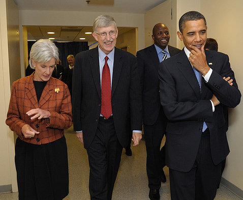 A smiling Collins walks down the hall flanked by then-HHS Secretary Sebelius and then-President Barack Obama who's holding his hand under in chin in thought.