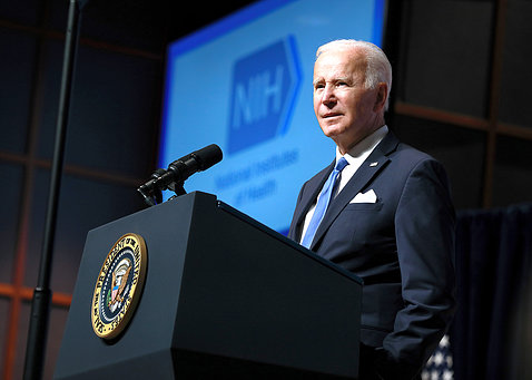 President Biden at NIH podium with NIH logo projected on slide behind him.