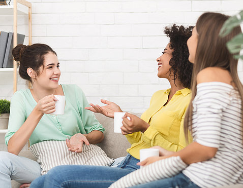 Three young women sit on a couch, drink coffee with each and laugh