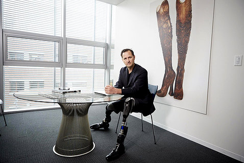 Herr is smiling, seated at a table. His prosthetic legs are prominent. On a wall behind him is an artwork of a pair of legs.