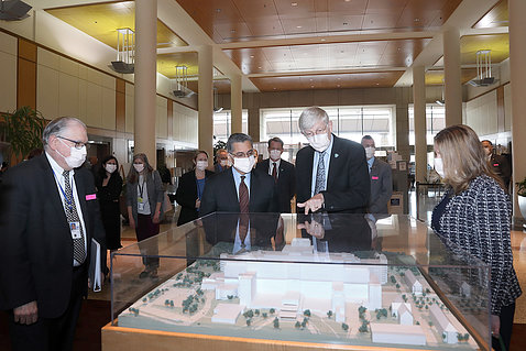 Four individuals in face masks gather around a large pedestal topped with an architectural model of a building under glass.
