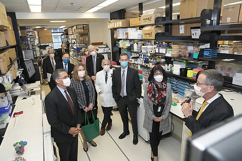 Several individuals in face masks gathered in a lab as one man speaks.