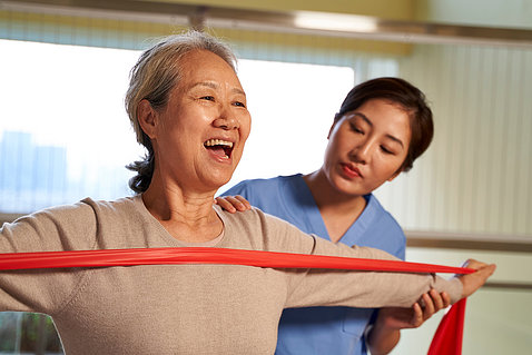 An older woman uses a theraband while a health care worker supervises