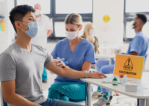 An Asian man in a grey t-shirt rests his arm on a table as nurse in blue scrubs holds up a needle. A yellow sign reading Covid Vaccination sits on the table.