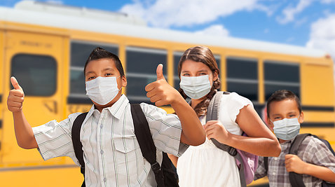 Three Latino kids, wearing backpacks and smiling behind their masks, give thumbs up - in front of a school bus with a bright blue sky overhead.