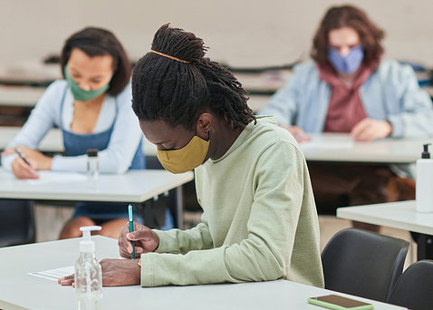 Three high school students wearing masks sit at school desks with pens in hand, bottles of hand sanitizer nearby. 