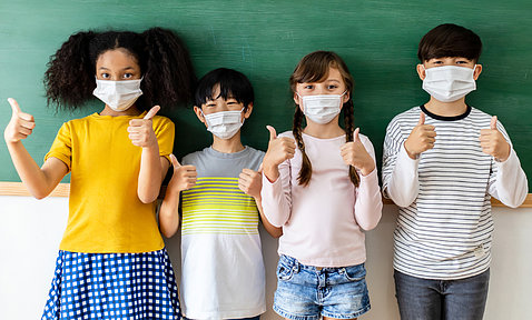 Four children wearing masks with thumbs up, standing in front of chalkboard