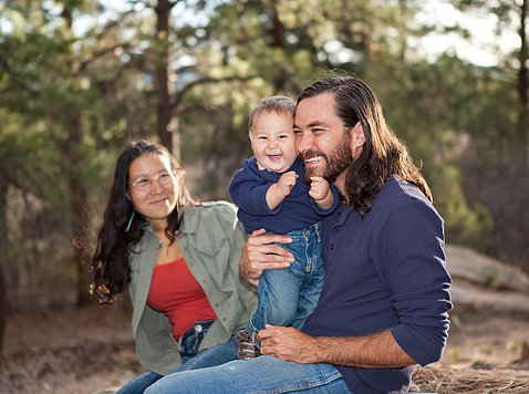 An American Indian couple hold their smiling baby outside, surrounded by trees.