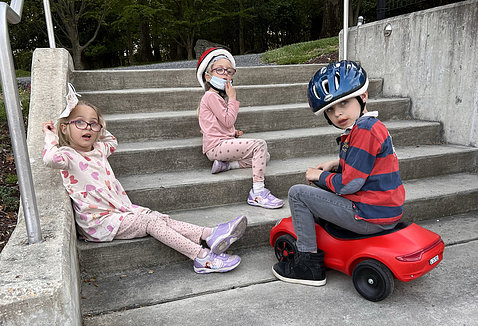 The twins wearing pink and Hampus sitting on a small red car play on the steps near the Children's Inn playground.