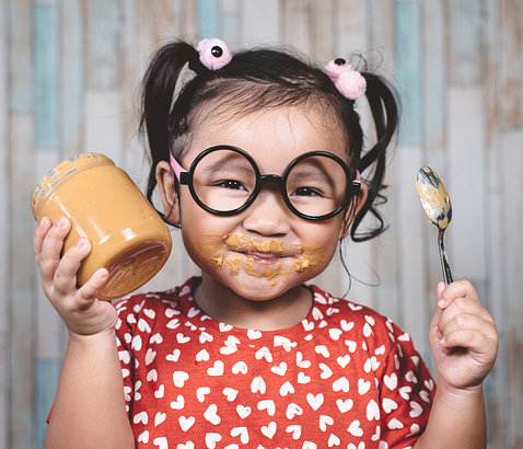 An Asian child in big round glasses and pigtails smiles as she holds up a jar of peanut butter and a spoon; her cheeks are smeared with peanut butter.