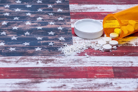 On a table with a fading painted American flag, a medicine bottle lies on its side out of which pours pills and powder.
