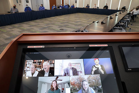 About a dozen staff members sit spaced, masked around a large conference table. A video monitor in the foreground shows event speakers.