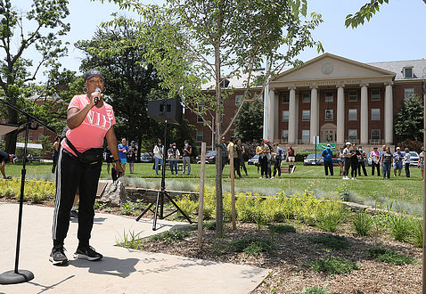 Edwards in pink NIH shirt talks into microphone as dozens of hikers stand in front of building 1 waiting for hike to begin.