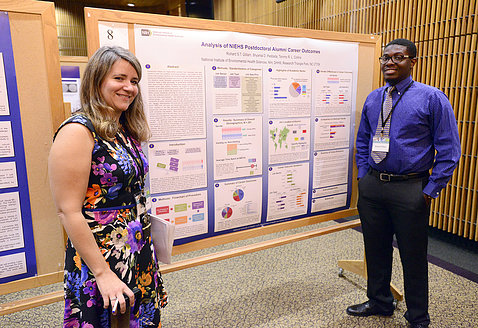 Collins and Gilliam stand smiling next to a colorful scientific poster titled: analysis of NIEHS postdoctoral alumni career outcomes