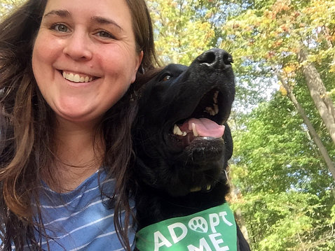 Connolly smiles with her black dog, who is wearing a green bandana with the words, "Adopt Me"