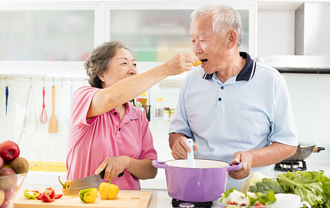An older Asian couple cooking in the kitchen; the woman is feeding the man while he holds a purple pot on the stove.