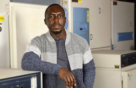 Dr. Adedeji stands with arm leaning on a centrifuge, surrounded by freezers and other lab equipment.