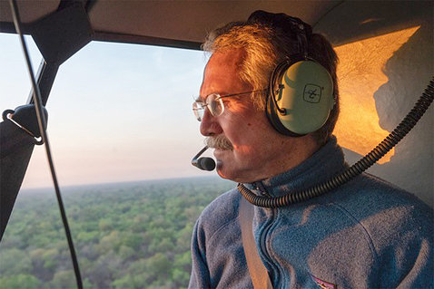 Quammen wearing an aviation headset sits in helicopter looking out; trees in view out side window