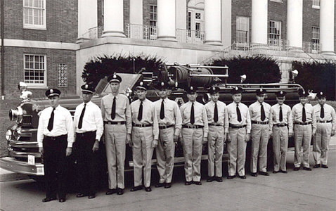 Black & white photo of fire department staff standing next to fire truck in front of Bldg. 1