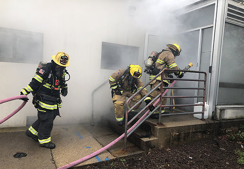 Three fire fighters in uniform walking into smoky building with hose on NIH campus, as part of a training exercise