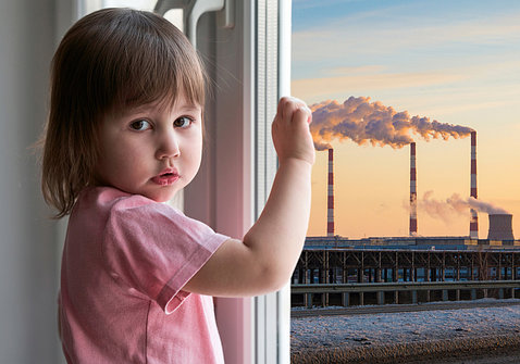 A young girl in a pink shirt holds hand to window, with view of smokestacks outside.