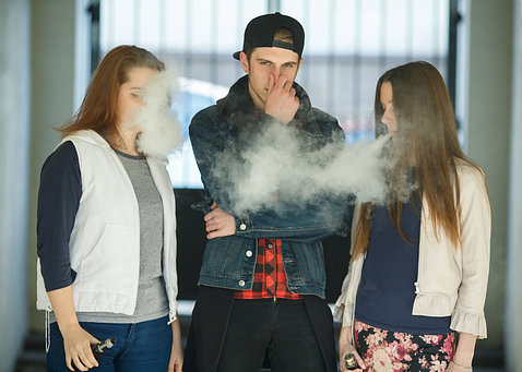Two teen girls blow smoke while vaping while teen boy in the middle abstains, holds his nose.