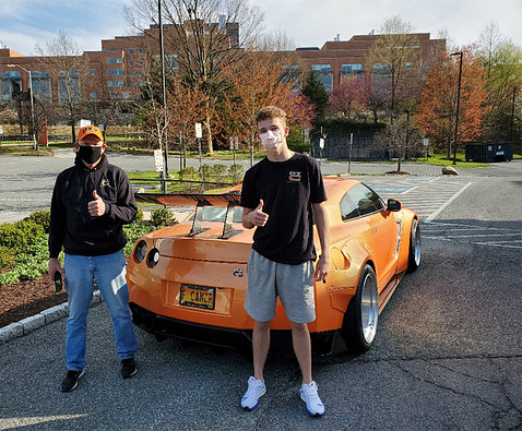 Bruce and patient Luke give thumbs up standing behind bright orange Driven to Cure sports car, parked behind the Clinical Center