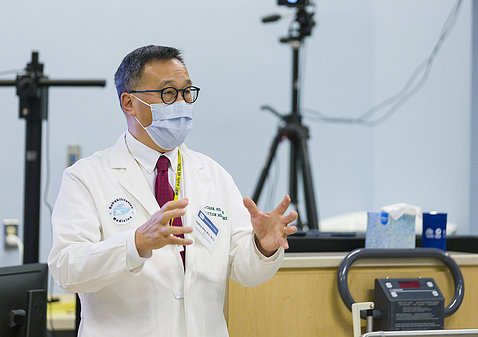 Dr. Chan, wearing white lab coat and holding hands out in front, discusses rehab at NIH.