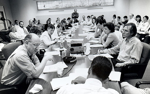 Black and white photo showing Rosenberg sitting and talking with others at long conference table in crowded room