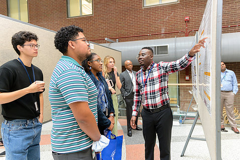 Man in plaid shirt explains his research poster to onlookers