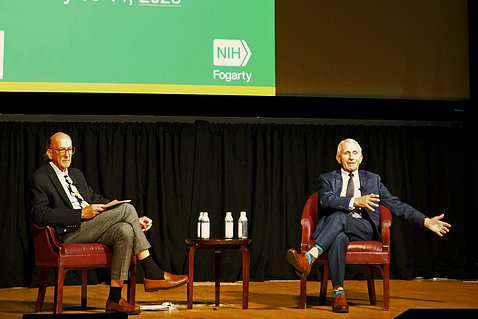 Glass, left, and Fauci, right, sit opposite each other on stage with a green slide show displayed behind them