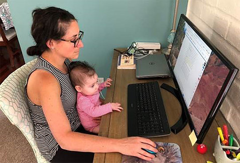 Gabriel sits by computer, hand on mouse, with her baby daughter on her lab - both looking at screen.
