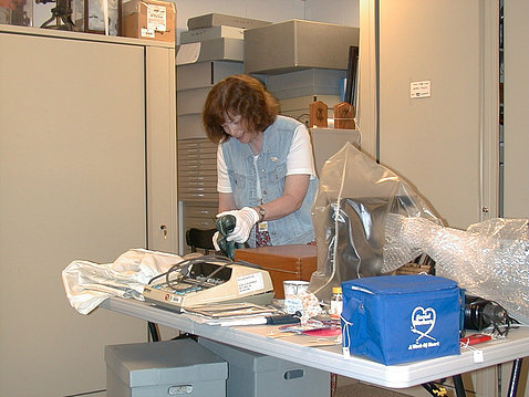 Lyons stands over a table crowded with objects. She wears white gloves as she examines an artifact.