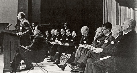 Black&white image of man standing at podium with several more men seated on stage behind him.