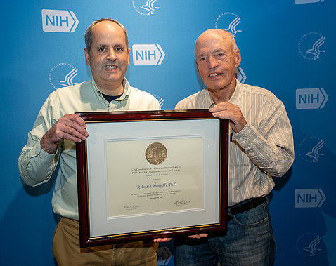 Lorsch and Young stand smiling with plaque, against blue NIH backdrop.