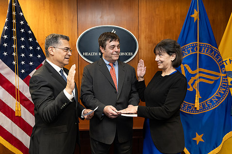 Becerra raise right hand, faces Monica Bertagnolli who has right hand raised. Bertagnolli's husband Alex holds a book, and smiles.