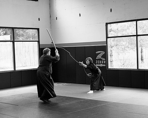 B&W photo of man in martial arts pants and tunic holds up rod facing woman in similar uniform, slightly crouching, holding up rod and rope. 