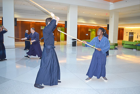In foreground, man and woman, both in wide-legged martial arts pants and tunic, face each other holding a sword and a long rod.