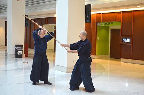 Two men in martial arts tunics and pants face off holding a sword and a rod.