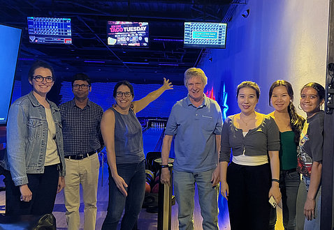 Samara, smiling, extends arm toward scoreboard screen in bowling alley, with lab colleagues by her side