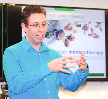 Green stands talking, cupping his hands, in front of immunotherapy poster.