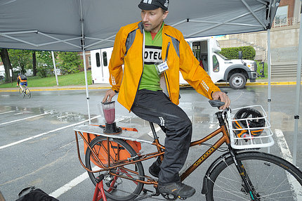 Under a tent, cyclist pedals bike to operate attached blender that is making smoothies.