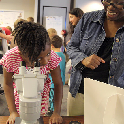 A young girl looks through a microscope as her mom looks on, smiling.