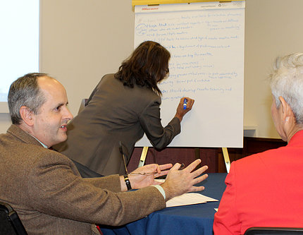 Seated Lorsch speaks to tablemate, with woman writing on presentation pad in background
