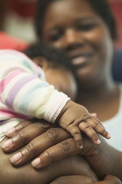 A smiling African American woman snuggles with her baby, whose little hand lays on top of hers.