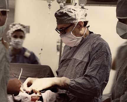 Tinted photo of masked and gowned Debakey with his gloved hands poised over an unseen patient