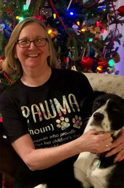 A smiling Jill in a black t-shirt that reads "PawMa" sits at home in front of a Christmas tree snuggled with black and white dog.