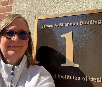 Jill George wearing sunglasses stands next to Shannon Bldg. plaque outside Bldg. 1.