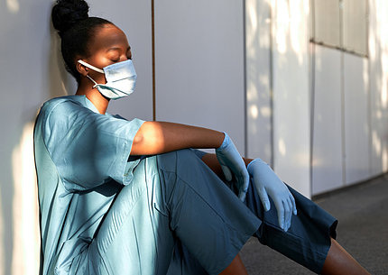 A female nurse in blue scrubs, mask and gloves sits, leaning against a hospital wall, asleep.