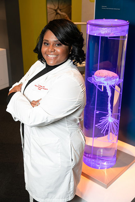 In lab coat, Jones stands with arms crossed, a model brain floating in an aquarium at her back.